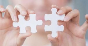 Hands holding two white puzzle pieces, almost connecting